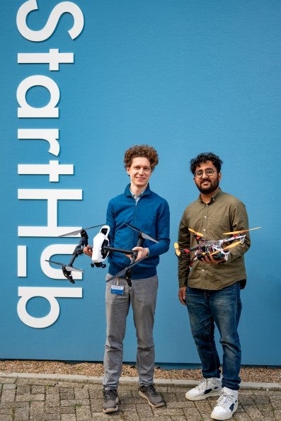 Aleksey Martynyuk and Aryan Sharma pose holding their 3D printed drones in front of the blue exterior of the VU StartHub