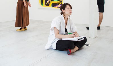  Woman sits on the floor in front of a work of art in a museum and makes notes in her sketch book.