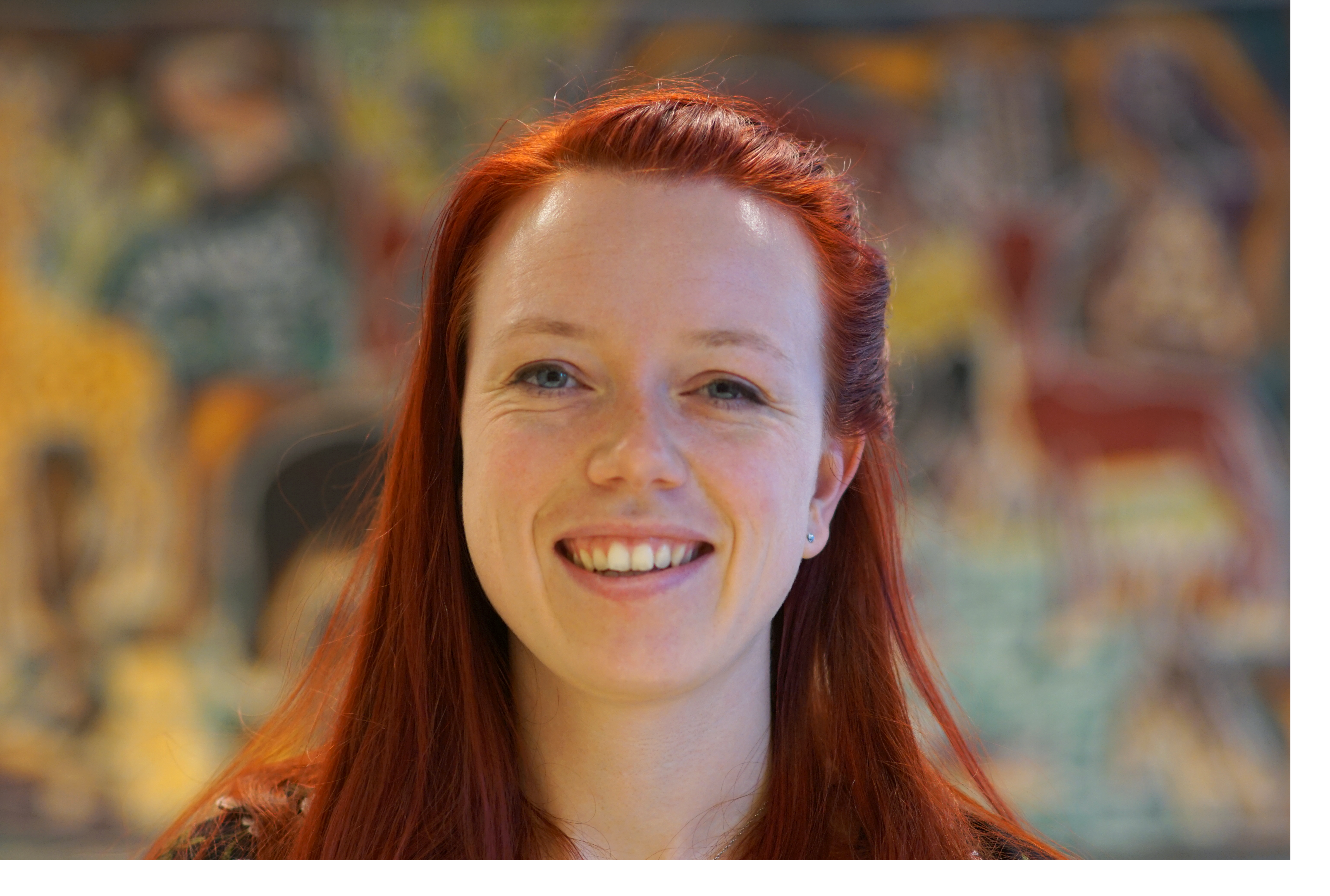 Broad-smiling woman with long red hair against a vaguely colourful background