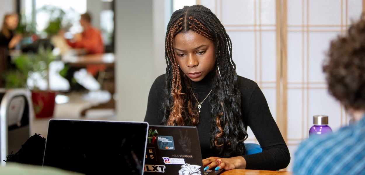 A young woman with braided hair is intently working on a laptop in an office environment.