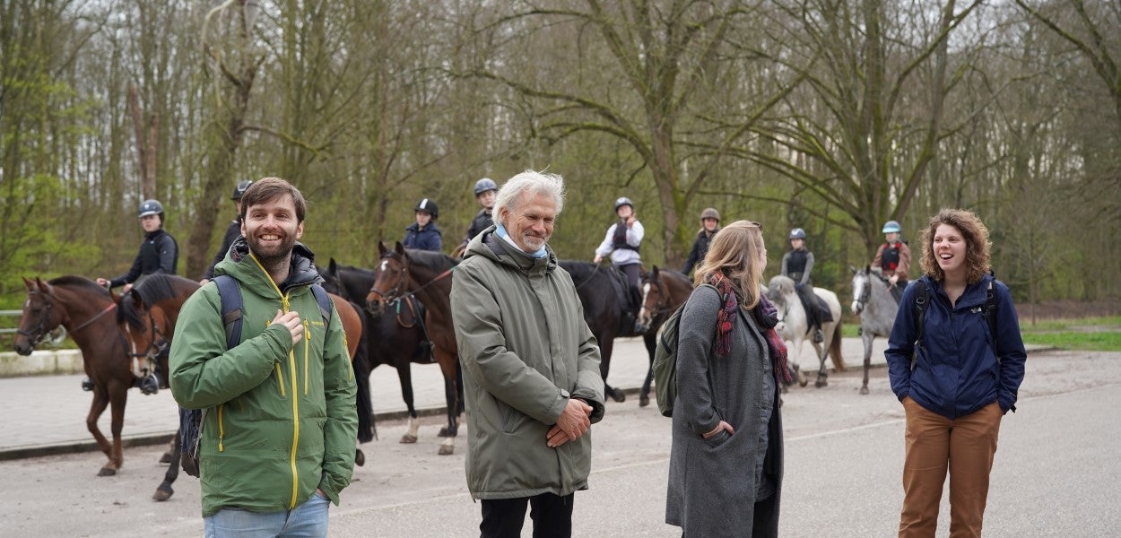 People are casually talking while standing on a path with a group of equestrians in the background.