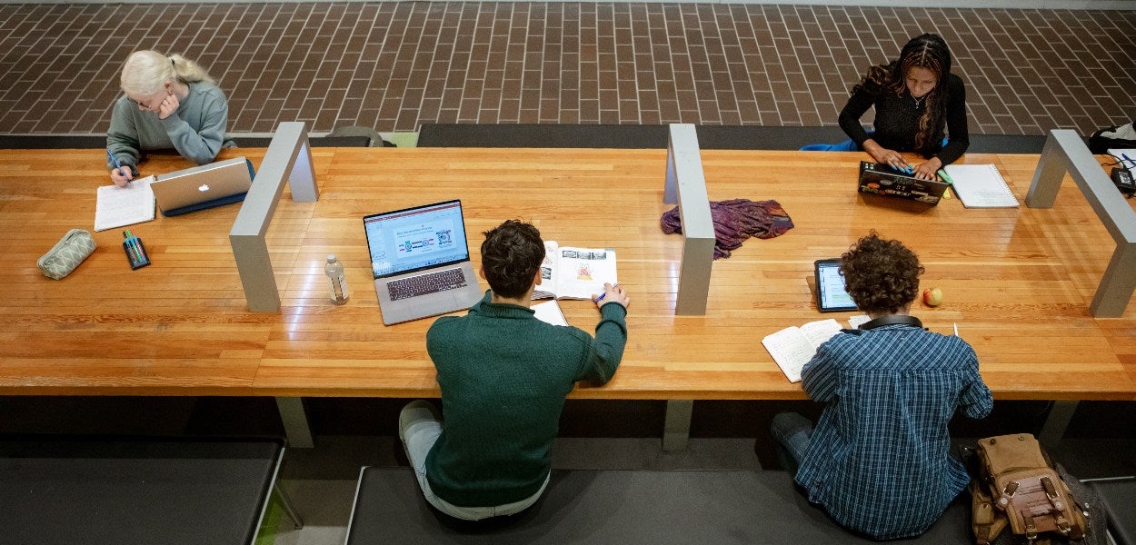 Four students studying at a long wooden table in a library, each with their own laptops and study materials.