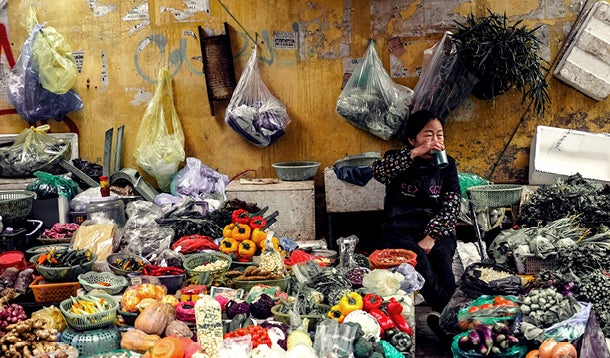 Woman drinks from her cup behind a fully packed market stall, she sells various foods