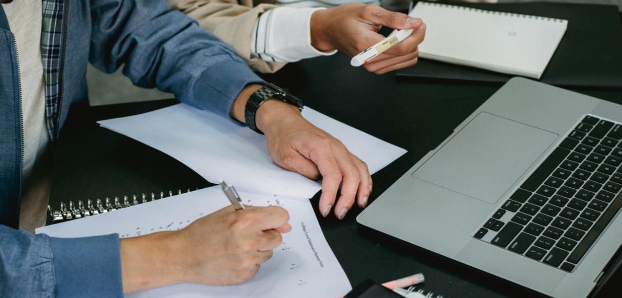 Twee mensen, de een in een blauw overhemd en de ander in een wit overhemd, hebben een pen in hun handen terwijl ze aan het werk zijn op een bureau waar een open laptop op ligt. 