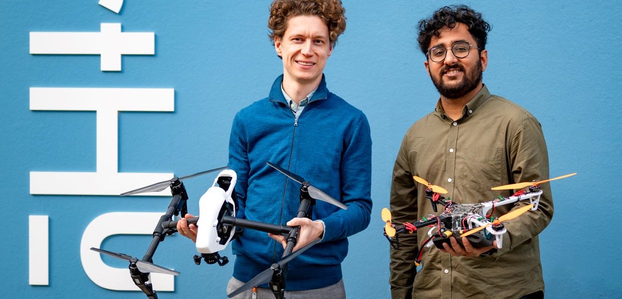 Aleksey Martynyuk en Aryan Sharma poseren met hun 3D-geprinte drones in de hand voor de hemelsblauwe buitenmuur van de VU StartHub