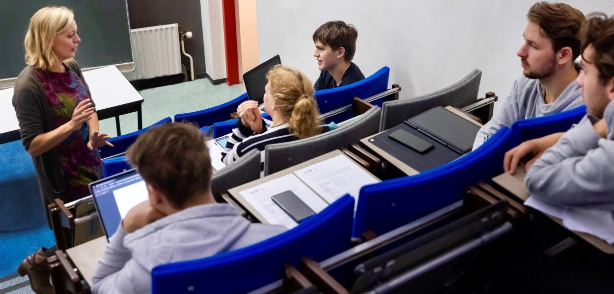 Docente legt een opdracht uit aan 5 aandachtig luisterende studenten in een collegezaal met blauwe stoelen