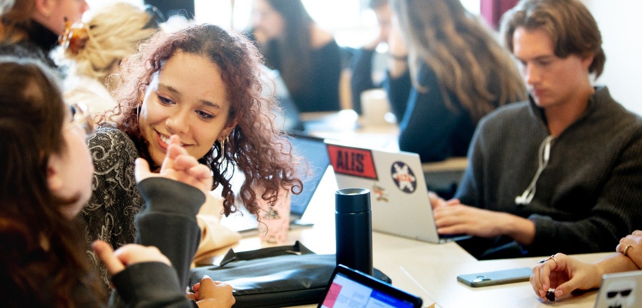 A girl with curly hair sitting in a room full of students working on their laptops, listens to her fellow student's story with a smile. 