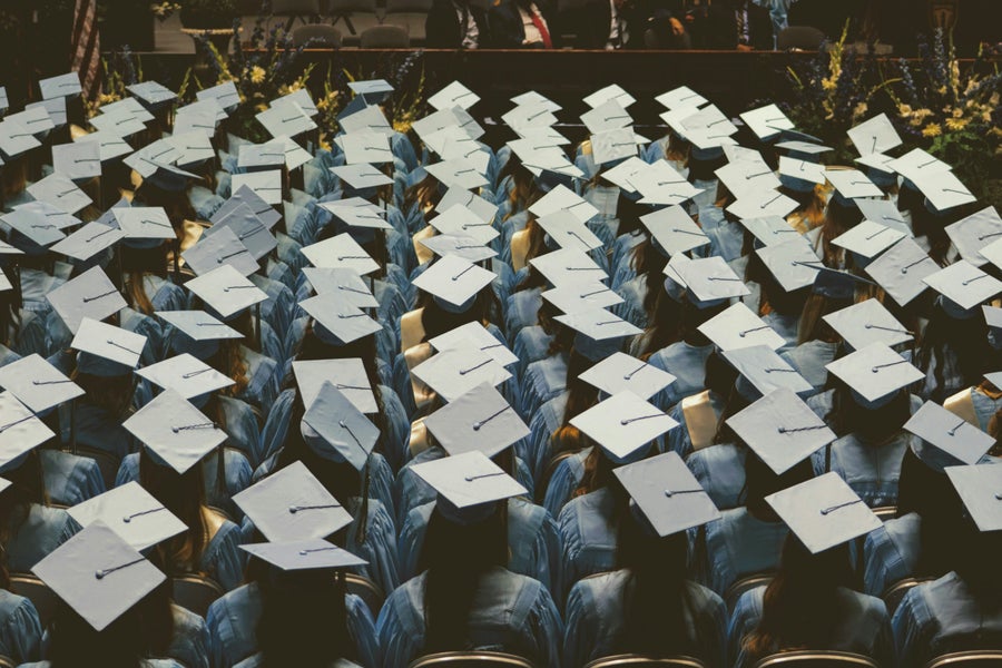 
Een groep afgestudeerden in afstudeertoga’s en -baretten zit klaar voor hun diploma-uitreiking, gezien vanaf de achterkant. 