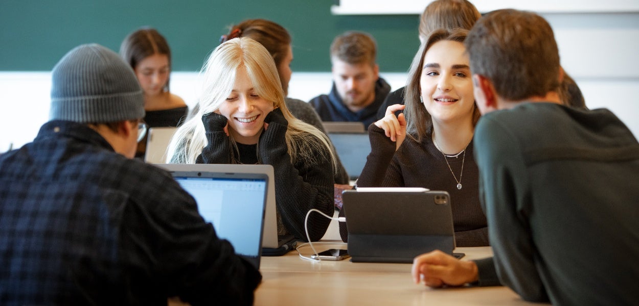 Studenten werken samen aan een tafel met laptops en tablets, terwijl ze glimlachen en met elkaar praten.