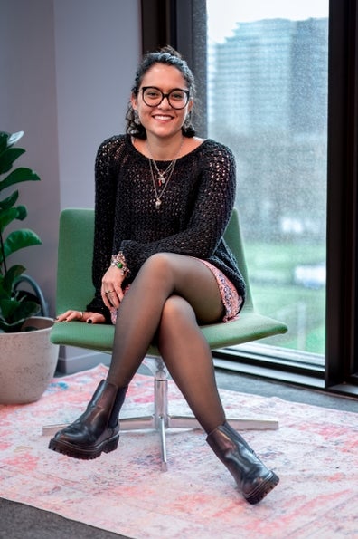Een vrouw met bril, gekleed in een zwarte outfit, zit glimlachend op een groene stoel in een kantooromgeving